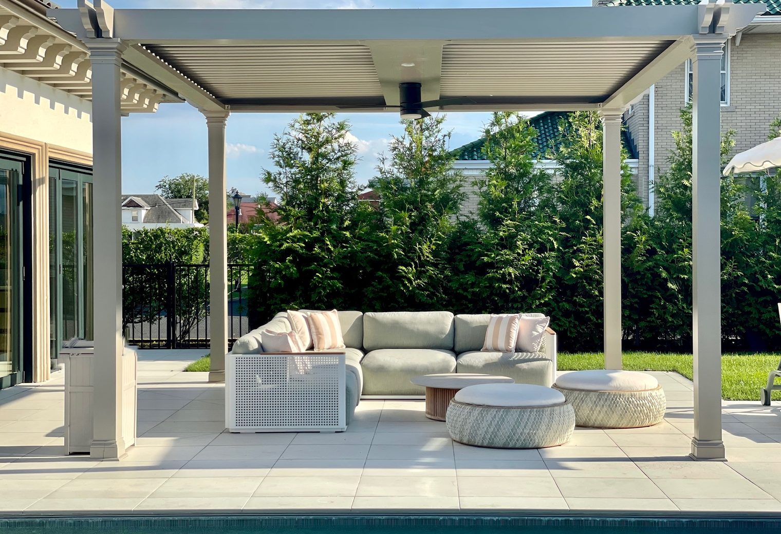 A louvered pergola over a sectional sofa and woven poufs beside the pool