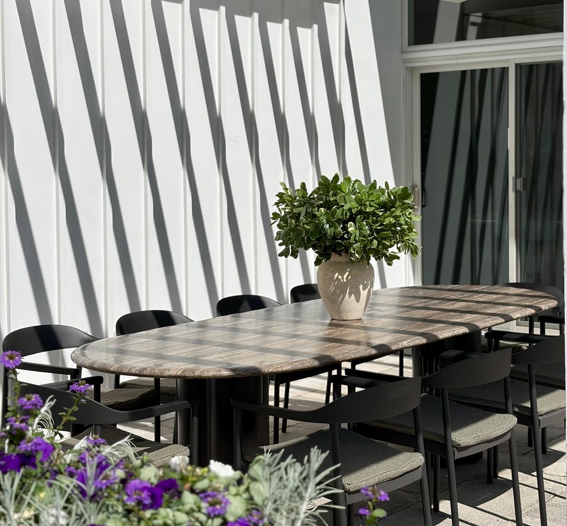 An oval travertine dining table with black chairs on a sunlit terrace